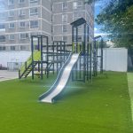 Outdoor play structure with various climbing features, slides, and multiple platforms in a playground setting, surrounded by trees and located near modern buildings.