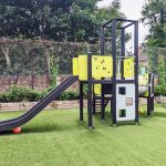 Outdoor play structure with a slide, climbing features, and multiple platforms in a playground setting, surrounded by greenery and a chain-link fence with trees in the background.