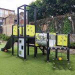 Outdoor play structure with a slide, climbing features, and multiple platforms in a playground setting, surrounded by greenery and a chain-link fence with trees in the background.
