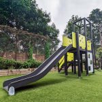 Outdoor play structure with a slide, climbing features, and multiple platforms in a playground setting, surrounded by greenery and a chain-link fence with trees in the background.