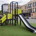 Outdoor play structure with slides, climbing features, and multiple platforms in a playground setting, located in front of a modern brick building with large windows and a play area with colourful markings.