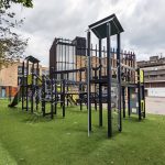 Outdoor play structure with various climbing features, slides, and multiple platforms in a playground setting, surrounded by trees and located near modern buildings.