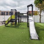 Outdoor play structure with various climbing features, slides, and multiple platforms in a playground setting, surrounded by trees and located near modern buildings.