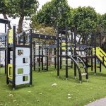Modern playground equipment featuring a combination of slides, climbing elements, and elevated play areas, situated on an artificial grass surface with trees and nearby buildings.