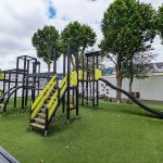 Complex outdoor play structure with a variety of climbing and sliding options, positioned on artificial turf in a playground area, with trees and modern buildings surrounding the space.